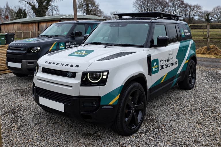 A white and a dark grey Land Rover Defender in Yorkshire 3D Scanning livery, parked on a gravel yard